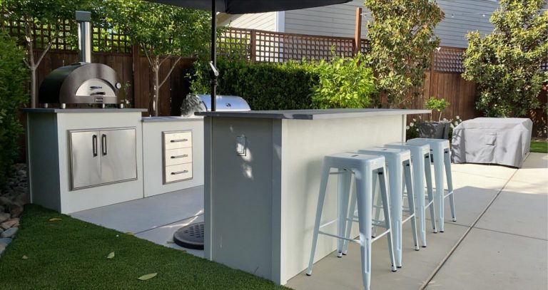 Modern outdoor kitchen setup with white cabinetry, stainless steel appliances, and blue bar stools in a backyard setting