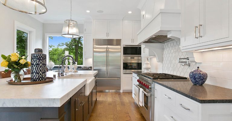 Modern white kitchen with stainless steel appliances, large island, marble countertops, and natural lighting from windows