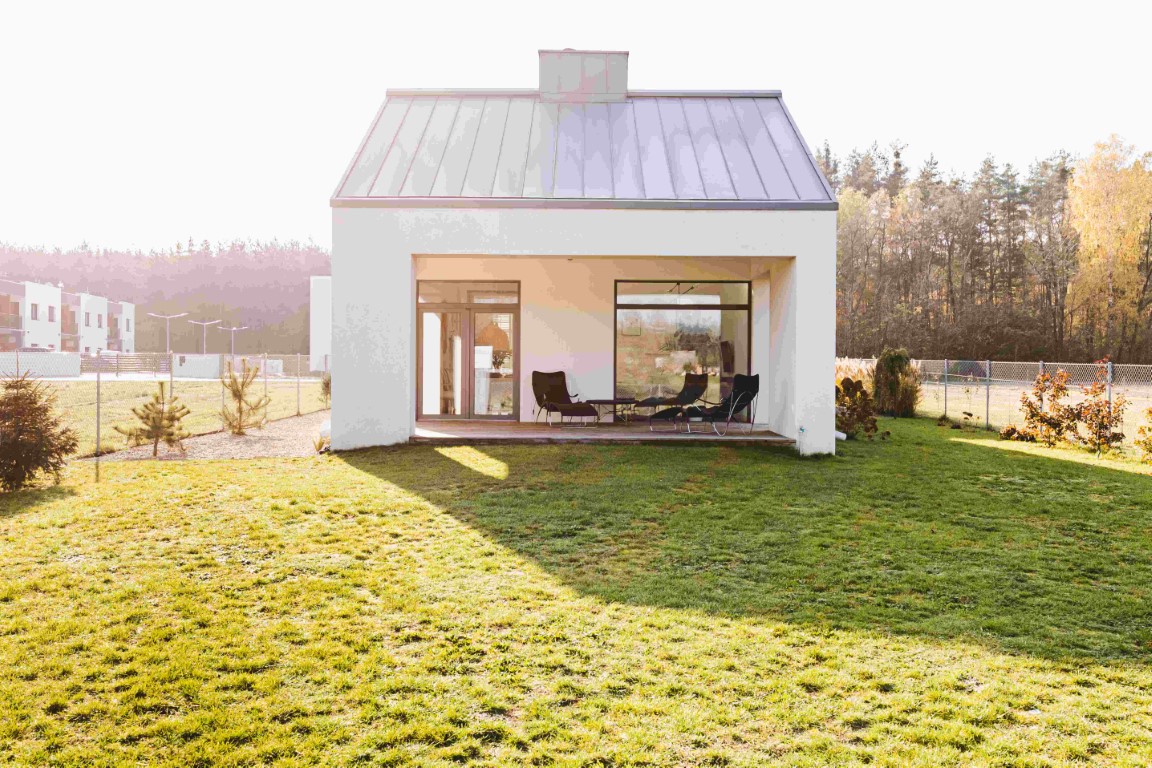 Modern small white house with large windows on grassy lawn surrounded by trees