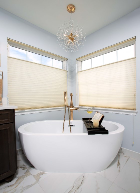 Modern white freestanding bathtub with chrome fixtures under a crystal chandelier and transom windows with cellular shades