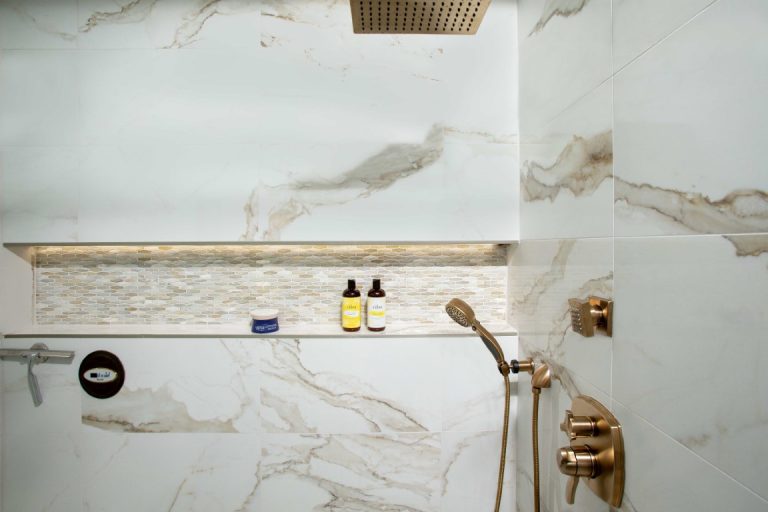 Modern bathroom shower wall with marble tile, brass fixtures, and organized toiletries on floating shelf