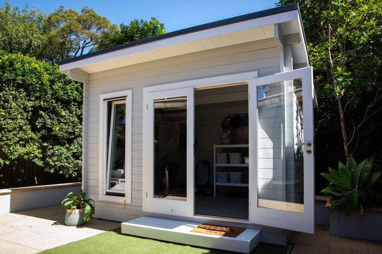 Modern white garden studio with glass doors, shelving, deck entrance, and surrounding greenery